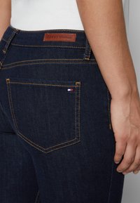 Mörkblå denimjeans med en läderetikett märkt "TOMMY HILFIGER" baktill, orange sömmar och en liten röd, vit och blå logotyp på fickan.