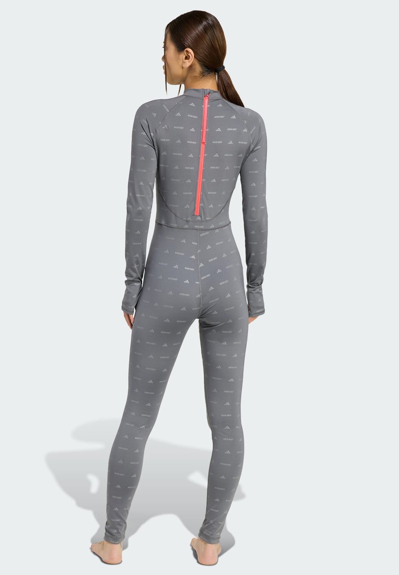 Vrouw die blootsvoets met de rug naar je toe staat, gekleed in een grijze overall met lange mouwen en een rode verticale rits op de rug.