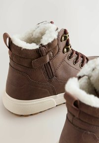 Braune Knöchelstiefel mit einem weichen, flauschigen Futter; verfügen über einen Reißverschluss und eine Schnürdesign. Der Hintergrund ist neutral und hebt das Schuhwerk hervor.