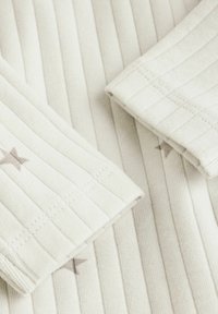 Lys beige stof med lodrette ribbede tekstur, der har subtile stjerne mønstre i en mørkere nuance. Foldede ærmer vises.