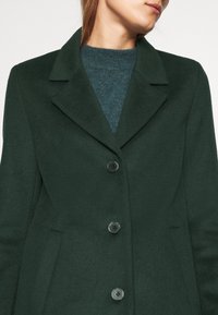 Cappotto di lana verde scuro con colletto a revers, cinque bottoni neri e tasche laterali. Indossato sopra un maglione blu-verdastro a trama.