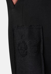 Schwarzer Stoff mit einem gestickten Patch, der ein erhabenes Oberflächendesign aufweist, das die Buchstaben "ge" in schwarzem Faden darstellt.