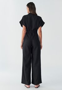 Mono negro con manga corta, cintura ceñida con cinturón, piernas anchas y textura lisa. La parte trasera cuenta con un cuello clásico y sin estampado.