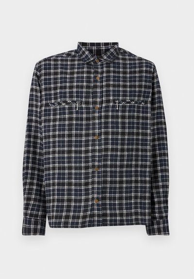 Camicia a scacchi in blu navy scuro con linee bianche. Presenta due tasche sul petto e bottoni marroni. Realizzata in un tessuto di cotone testurizzato.
