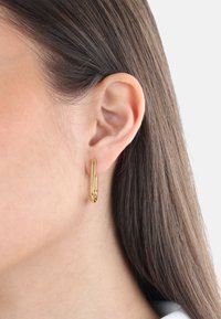 Pendiente de aro en tono dorado, forma ovalada alargada, superficie lisa, diseño minimalista. Lleva puesto en la oreja de un modelo con el cabello liso cerca.