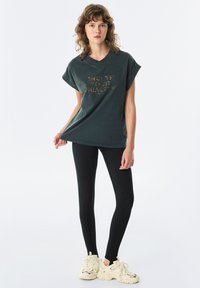 Ciemnozielony, oversize t-shirt z krótkimi rękawami, z teksturowaną złotą grafiką. Połączony z czarnymi legginsami i kremowymi butami sportowymi.