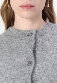 Grå strikket cardigan med rund hals, to grå knapper og myk tekstur. Stoffet viser subtile variasjoner i farge.