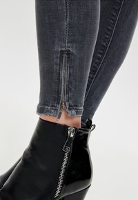 Primer plano de piernas inferiores que llevan jeans skinny gris oscuro con cremalleras en los tobillos y botas de cuero negras con cremalleras plateadas.