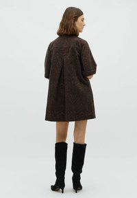 Eine Frau, die seitlich steht und ein lockeres, dunkelbraunes Kleid mit Tiermuster und Ellenbogenlangen Ärmeln trägt. Sie hat schwarze Stiefeletten mit Absatz, die bis zur Mitte der Waden reichen.