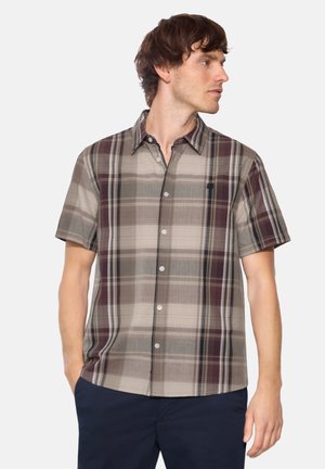 Junger Mann mit kurzärmligem kariertem Hemd in Beige-, Braun- und Grautönen, der nach rechts schaut und die Hände in den Taschen hat.