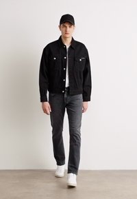Črna jeans jakna s srebrnimi gumbi, bela majica pod njo, sivi kavbojki in bele superge. Model nosi črno kapo.