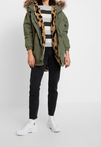 Green parka jacket with faux fur lining in leopard print, zip closure, adjustable hood, worn over a striped sweater and black jeans, paired with white boots.