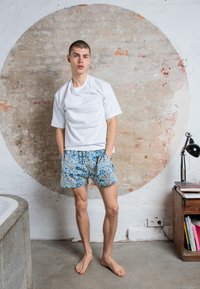 T-shirt bianca a maniche corte abbinata a pantaloni corti a fantasia blu con motivi floreali gialli e bianchi. A piedi nudi, in piedi contro un muro texture.