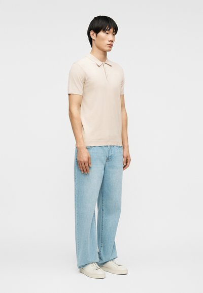 Homme debout portant un polo à manches courtes beige, un jean large bleu clair et des baskets blanches devant un fond uni.