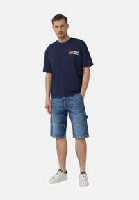 Navyblaues T-Shirt mit Logo, kombiniert mit blauen Denim-Cargo-Shorts bis zum Knie. Weiße Sneakers runden das Outfit ab.