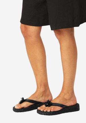 Ben iført sorte vævede platforms-flipflops med blomsterdekorationer på stropperne, kombineret med sorte knælange shorts.