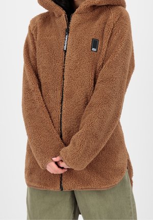 Mujer con chaqueta de forro polar sherpa marrón con cremallera y capucha, cremallera negra y pequeño parche negro con logo, combinada con pantalones verdes.