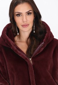 Burgundská kapucňová bunda vyrobená z plush materiálu, s uzatváraním na zips a jemnou textúrou. Ozdobená lesklou podšívkou a plush lemovaním.