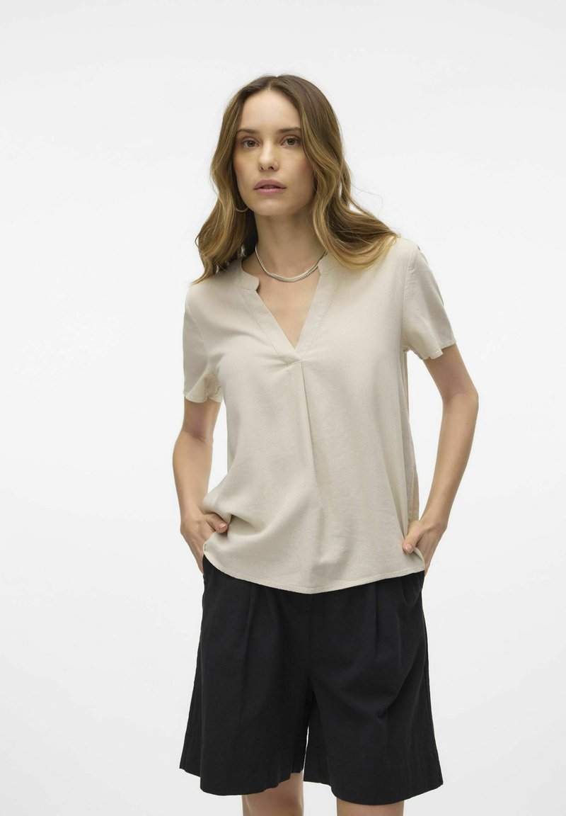 Top beige de manga corta con escote en V y detalle de pliegues, combinado con pantalones cortos negros de cintura alta, que presentan un ajuste relajado y una textura suave.