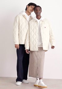 Giubbotti trapuntati beige chiaro indossati sopra felpe, abbinati a una gonna maxi beige e pantaloni scuri. Sneakers bianche con dettagli in gomma.
