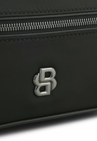 Borsa di stoffa nera con una cerniera argentata e un logo metallico. Il materiale è liscio con una finitura testurizzata attorno all'area della cerniera.