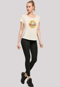 Cream t-shirt with a colorful Guns N' Roses graphic, short sleeves, and a relaxed fit, paired with black leggings and sneakers.