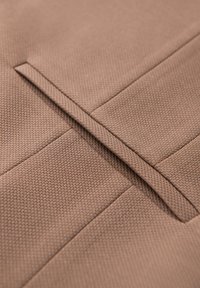 Gros plan d'un tissu marron texturé avec une fente rectangulaire horizontale cousue, probablement un détail d'ouverture de poche.