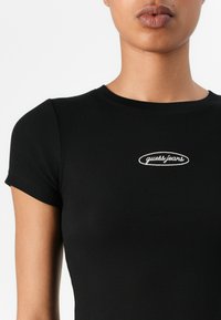 Černé žebrované tričko s přiléhavým střihem, krátkými rukávy a kulatým výstřihem. Na hrudi je vyšitý bílý logo "guess.jeans".