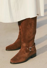 Cizme din suede maro, până la genunchi, cu toc mic și curele cu cataramă pe lateral, purtate cu o fustă plisată albă pe un pavement deschis.