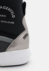 Zblízka na černý a metalický podpatek tenisky s bílou podrážkou, s kovovým logem "KARL" a vyšitým textem "KARL LAGERFELD PARIS" na textilním svršku.