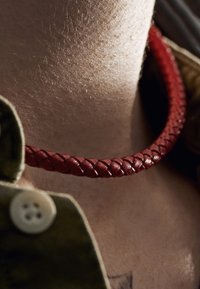 Ett rött flätat läderhalsband vilar mot huden, kompletterat med en grön tygkrage med en synlig knapp. Slät textur och detaljerad tajt vävning.