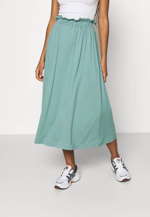 Femme portant une jupe midi vert turquoise clair avec une taille élastique froncée et des baskets blanches, debout devant un fond blanc.