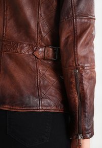 Giacca di pelle marrone con motivo trapuntato, vita con cintura e maniche con zip. Texture liscia con una leggera lucentezza.