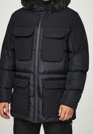 Veste d'hiver matelassée noire avec quatre poches à rabat à l'avant, fermeture éclair et capuche doublée de fourrure portée par une personne debout.