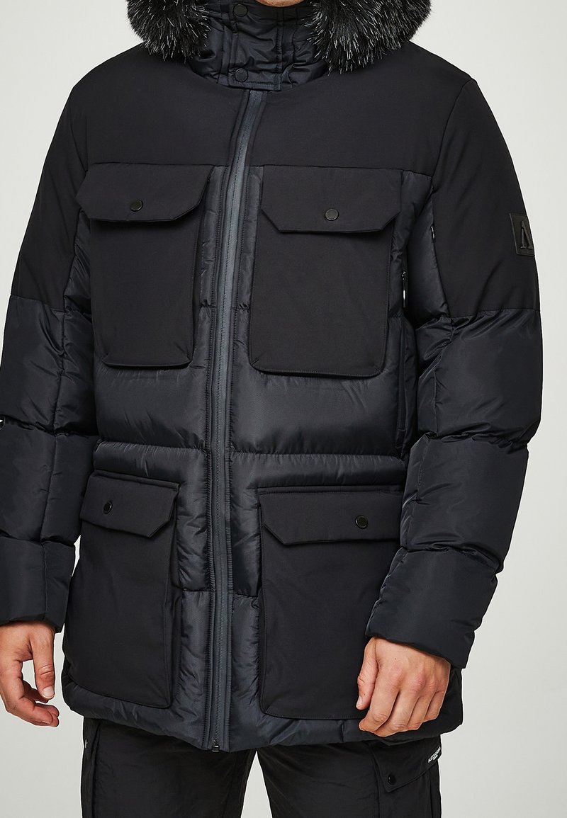 Veste d'hiver matelassée noire avec quatre poches à rabat à l'avant, fermeture éclair et capuche doublée de fourrure portée par une personne debout.