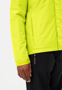 Limoengroene waterdichte jas met een gladde textuur, lange mouwen en klittenbandsluitingen, gecombineerd met zwarte broeken met een merklogo.