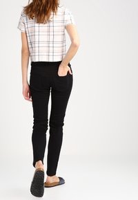 Witte cropped top met zwart ruitpatroon, zwarte skinny jeans en bruine sliders. Haar is los. Focus op de stof en pasvorm.