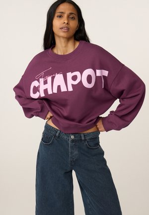 Vrouw draagt een paarse trui met de tekst "CHAPOT" en donkerblauwe spijkerbroek, staande met handen in de zij tegen een effen achtergrond.