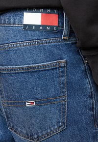 Bakfickan på blå denimjeans med Tommy Jeans logotypapplikation och liten flaggemblem, buren med en svart topp.