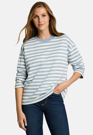 Vrouw met lang haar die een lichtblauw-wit gestreept shirt met lange mouwen en donkerblauwe spijkerbroek draagt, staand met één hand in haar zak.