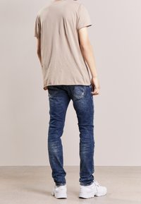 Lyhythihainen vaalean beige t-paita, jota on yhdistetty tummansinisiin farkkuihin, joissa on kulutuspintaa, ja valkoisiin lenkkareihin, nähtynä takaa.