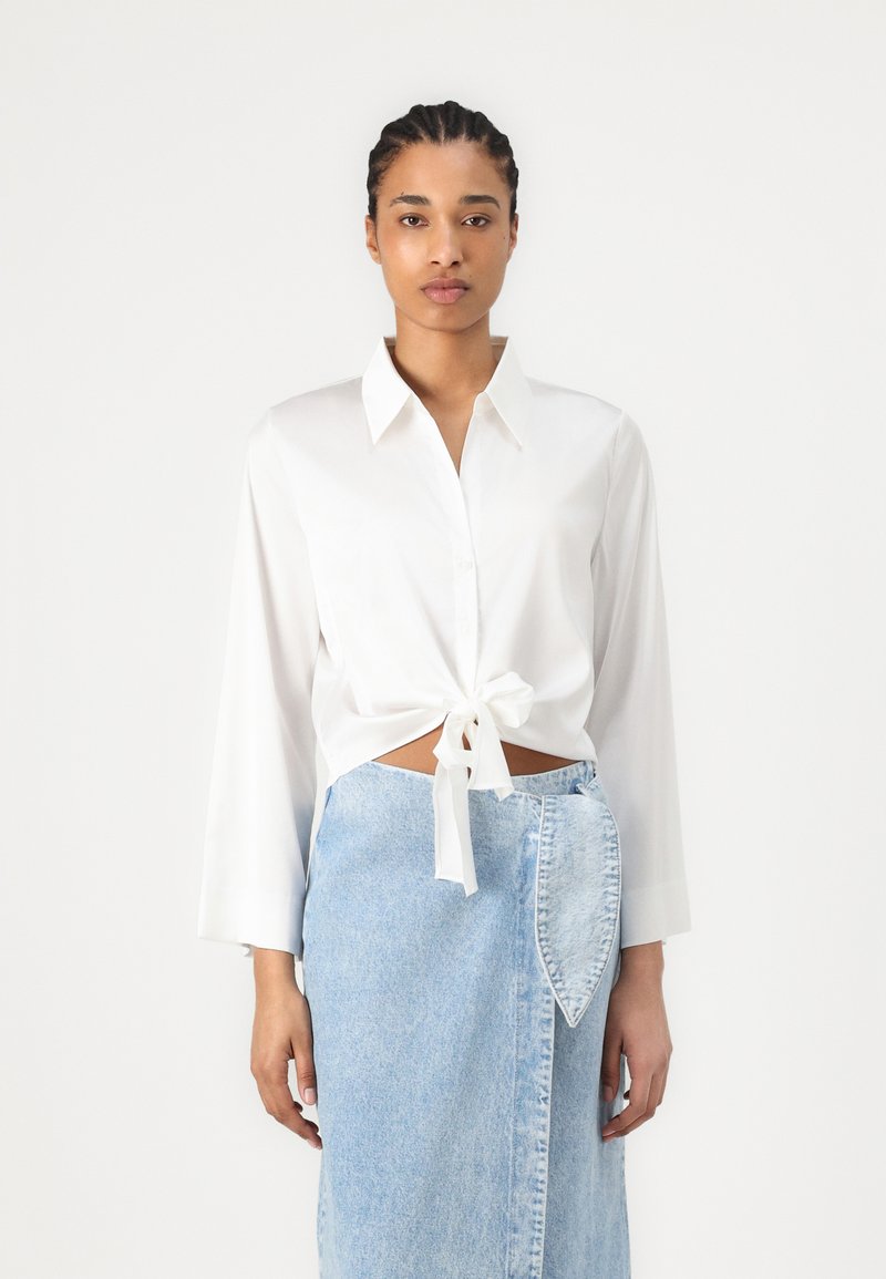 Camicia bianca con maniche lunghe, annodata in vita. Abbinata a una gonna di jeans azzurri chiari con una tasca e un taglio diagonale.