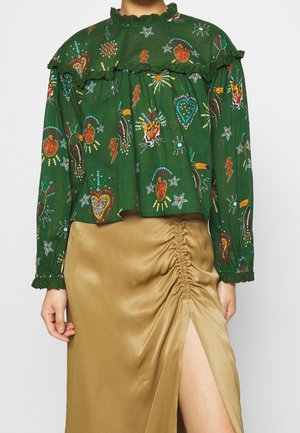 Grøn bluse med lange ærmer og farverige broderede motiver kombineret med en beige rynket nederdel med et højt slid.