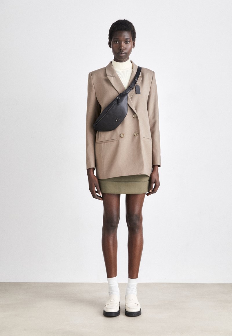 Beige dubbelgeknoopte blazer over een witte coltrui, gecombineerd met een olijfgroene minirok. Zwarte schoudertas, witte sokken en chunky witte schoenen.