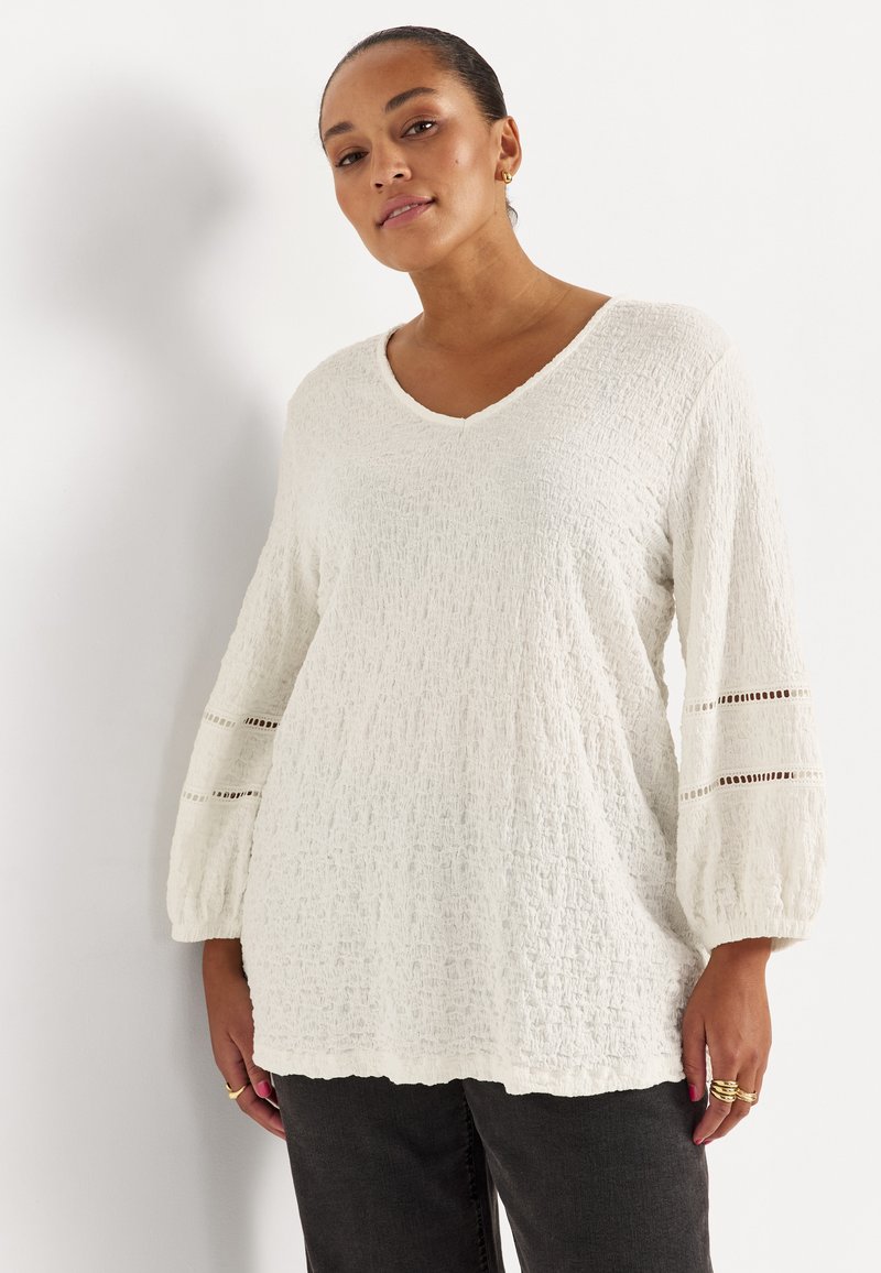 Frau trägt eine strukturierte weiße V-Ausschnitt-Bluse mit Ballonärmeln, die zwei horizontale Spitzen-Einsätze aufweist, kombiniert mit dunkler Hose.