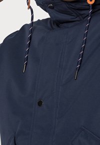 Marinblå jacka med hög krage, tryckknappar och en dragsko i huvan med kontrasterande detaljer och textilstruktur.