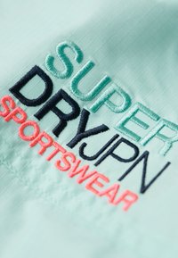 Broderi på lyseblåt stof med "SUPER DRY JPN" i grøn og sort og "SPORTSWEAR" i koralrosa tråd.