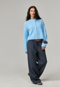 Cardigan tricotado azul claro, cortado, com fecho de botão. Emparelhado com calças largas navy e riscas, com cintura elástica.