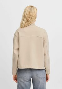 Giacca beige con un tessuto morbido e strutturato, vestibilità rilassata, colletto a coste e dettagli di cucitura sulla parte posteriore. Indossata con jeans grigi chiari.