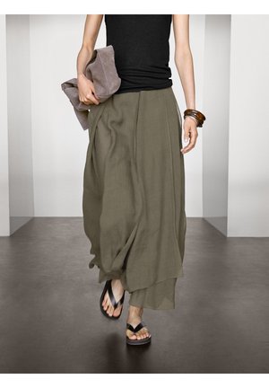 Jupe fluide vert olive, ample avec des plis ; associée à un débardeur noir, des tongs en plastique et une pochette gris clair.